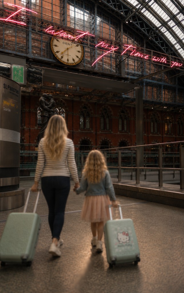 A parent and child walk hand in hand through St Pancras station, pulling suitcases beneath a pink neon sign that reads I want my Time with you.